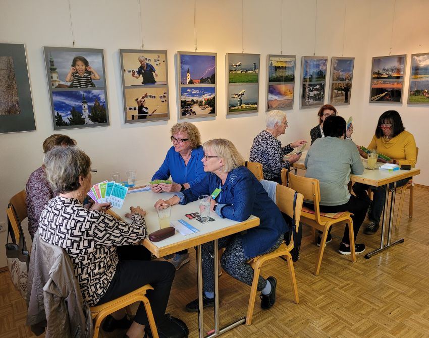 Je vier Senior:innen sitzen an zwei Tischen, Karten in der Handj, hinter ihnen Bilder an der Wand.