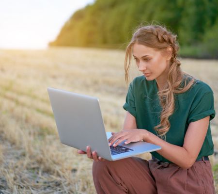 Frau mit grüner Bluse und herbstroter Plunderhose hock an einem abgemähtem Weizenfeld, im Hintergrund Wald. Sie hat einen geöffneten Laptop in der Hand und ihre Hand auf der Tastatur.