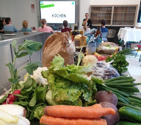Salat, Frühlingszwiebel, Käferbohnen und weiteres Gemüse liegen auf einer Küchenzeile. Im Hintergrund eine Person im Stehen auf die sitzenden Personen schauen