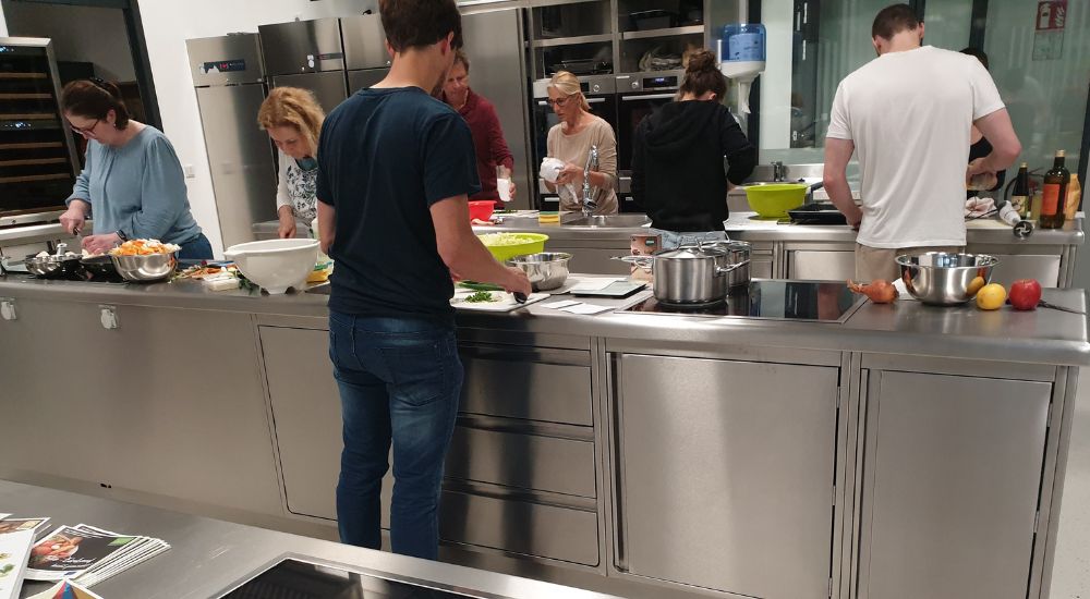 6 Personen beim Kochen rechts und links von zwei Kochzeilen stehend
