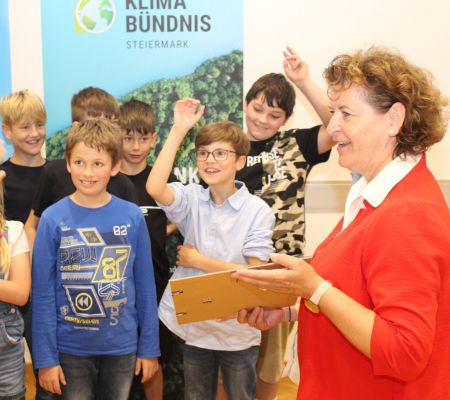 Frau im Vordergrund in rotem Oberteil mit Blatt Papier in der Hand, im Hintergrund Kinder, dahinter ein Roll-Up des Klimabündnis Steiermark