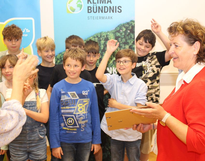 Frau im Vordergrund in rotem Oberteil mit Blatt Papier in der Hand, im Hintergrund Kinder, dahinter ein Roll-Up des Klimabündnis Steiermark