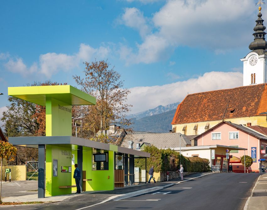 moderne Bushaltestelle in Grün gehalten, im Hintergrund eine Kirche